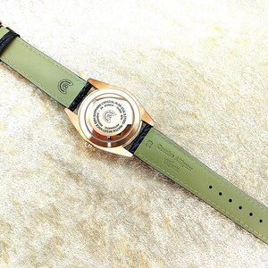 May include: A rose gold wristwatch with a black leather strap and a light green alligator leather strap. The watch face is visible, displaying text and intricate details. The watch band is approximately 2 cm wide.