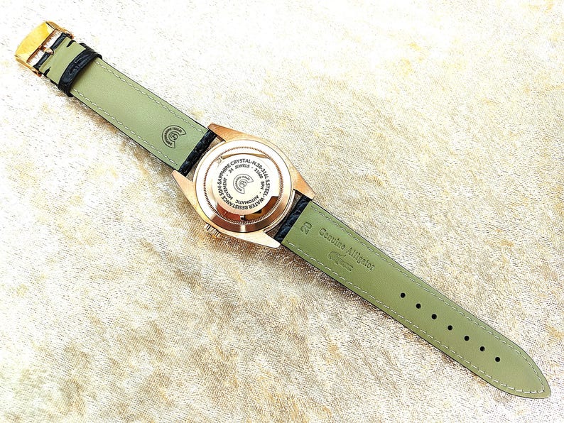 May include: A rose gold wristwatch with a black leather strap and a light green alligator leather strap. The watch face is visible, displaying text and intricate details. The watch band is approximately 2 cm wide.