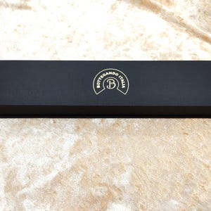 May include: A rectangular black box with a textured surface. The box features a gold emblem reading "Bottegando Italia" with a stylized "B". The box is on a beige surface.