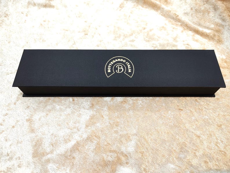 May include: A rectangular black box with a textured surface. The box features a gold emblem reading "Bottegando Italia" with a stylized "B". The box is on a beige surface.