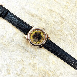 May include: A gold wristwatch with a black leather strap. The watch face has a yellow and black design, with visible internal mechanisms. The strap has a crocodile pattern and a gold buckle.