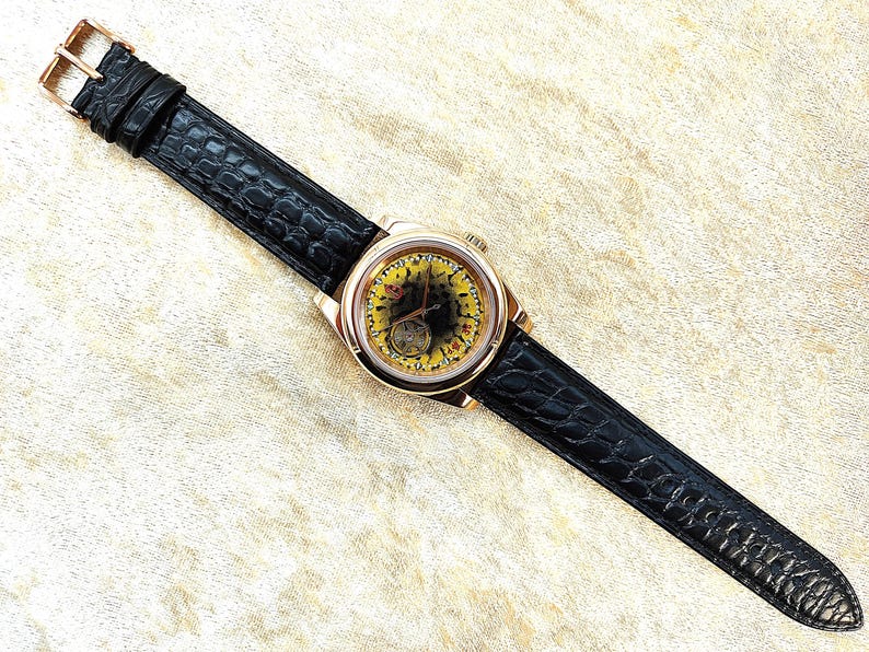 May include: A gold wristwatch with a black leather strap. The watch face has a yellow and black design, with visible internal mechanisms. The strap has a crocodile pattern and a gold buckle.