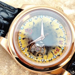 May include: A gold-coloured wristwatch with a black leather strap. The watch face has an intricate design with a visible mechanism, gold and yellow accents, and diamond-shaped markers. The hands are rose-gold coloured.