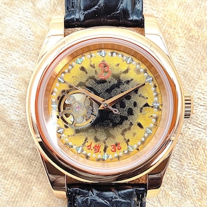 May include: A gold-coloured wristwatch with a black leather strap. The watch face has a yellow and black design, a visible mechanical movement, and gold hands. The watch has a round face and a textured dial.