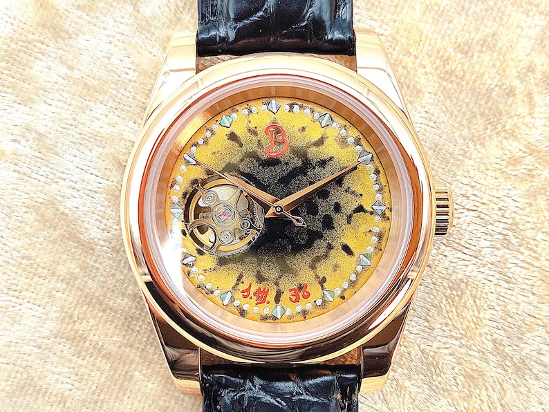 May include: A gold-coloured wristwatch with a black leather strap. The watch face has a yellow and black design, a visible mechanical movement, and gold hands. The watch has a round face and a textured dial.