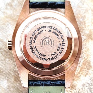 May include: Close-up of the back of a rose gold wristwatch with a black and green leather strap. The watch face has the text "50M-SAPPHIRE CRYSTAL" and "24 JEWELS". The watch is labeled "BOTTEGANDO ITALIA".