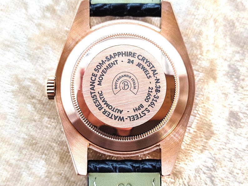 May include: Close-up of the back of a rose gold wristwatch with a black and green leather strap. The watch face has the text "50M-SAPPHIRE CRYSTAL" and "24 JEWELS". The watch is labeled "BOTTEGANDO ITALIA".
