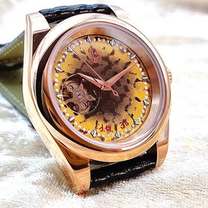 May include: A rose gold wristwatch with a yellow and brown patterned face, featuring a visible mechanical movement. The watch has rose gold hands and a black leather strap. The watch face has small diamond-shaped accents.