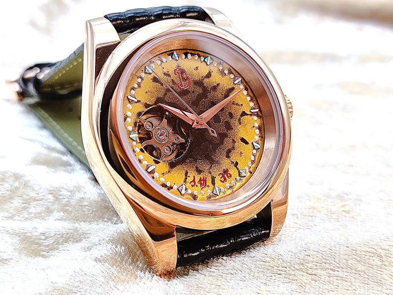 May include: A rose gold wristwatch with a yellow and brown patterned face, featuring a visible mechanical movement. The watch has rose gold hands and a black leather strap. The watch face has small diamond-shaped accents.