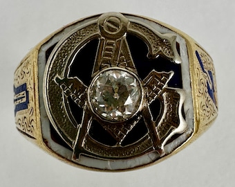 Puede incluir: Un anillo de oro con un diamante central y un emblema masónico. El anillo tiene una banda de oro con detalles de esmalte blanco y azul. El símbolo masónico incluye una brújula, una escuadra y la letra "G".