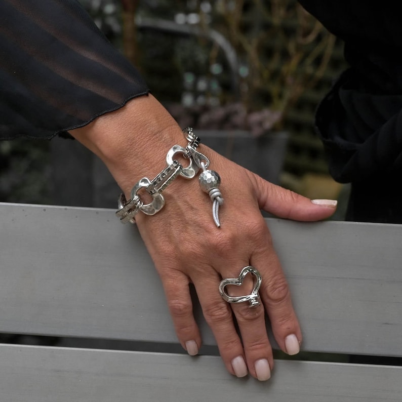 May include: A silver bracelet with a hammered metal design and a dangling charm. The bracelet is worn on a person's wrist. The person is also wearing a silver heart-shaped ring.