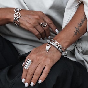 Op de afbeelding: Een hand draagt een zilveren ring en twee zilveren armbanden. De armband op de pols heeft een hartvormige hanger. De andere hand draagt een zilveren armband met een groot, chunky ontwerp. De persoon heeft een tatoeage op zijn arm die "be still" zegt.