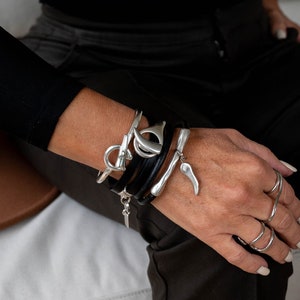 Peut inclure: Un bracelet en argent avec un bracelet en cuir et un charme en argent. Le bracelet est porté au poignet d'une personne.