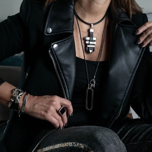 May include: A person wearing a black leather jacket with a silver and black layered pendant necklace and a silver ring. The necklace has a long chain with a silver oval pendant and a smaller silver charm.