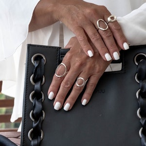 Peut inclure: Plusieurs bagues en argent aux formes ouvertes originales. Les doigts, vernis de blanc, portent les bijoux. Un sac à main noir, orné d'œillets argentés et d'une tresse noire, sert de support. Une blouse blanche est en arrière-plan.
