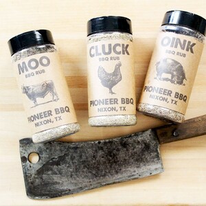 May include: Three spice jars labeled "MOO," "CLUCK," and "OINK" with animal illustrations, and the text "Pioneer BBQ Nixon, TX." A vintage meat cleaver rests below the jars on a wooden surface.