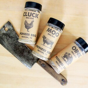 May include: Three spice rub jars labeled "Cluck," "Moo," and "Oink" with animal illustrations, and a cleaver on a wooden surface. The jars are from Pioneer BBQ, Nixon, TX. The jars have black lids and brown labels.