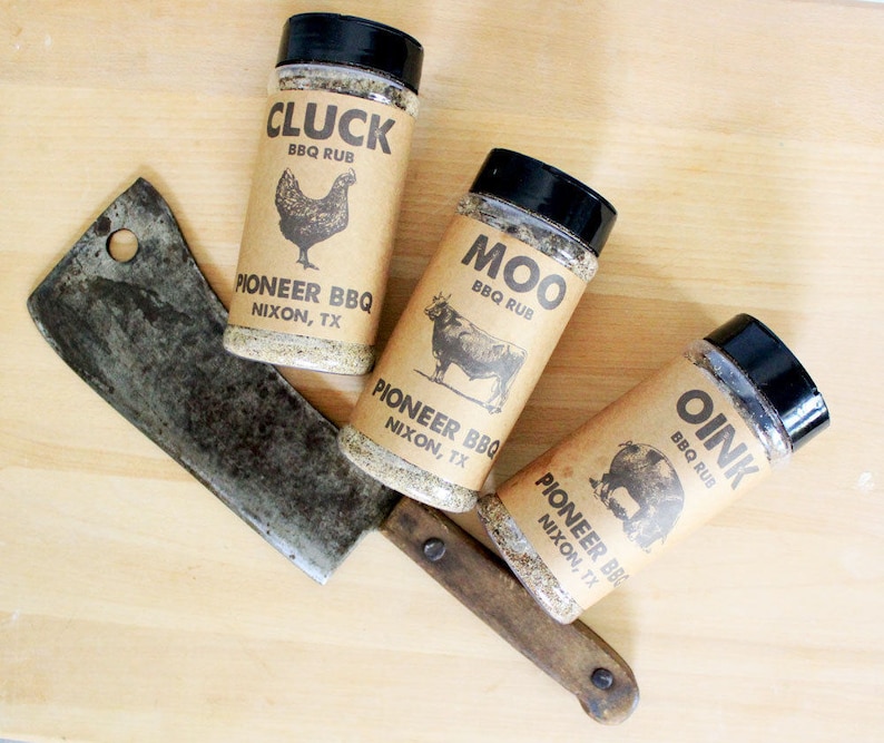 May include: Three spice rub jars labeled "Cluck," "Moo," and "Oink" with animal illustrations, and a cleaver on a wooden surface. The jars are from Pioneer BBQ, Nixon, TX. The jars have black lids and brown labels.