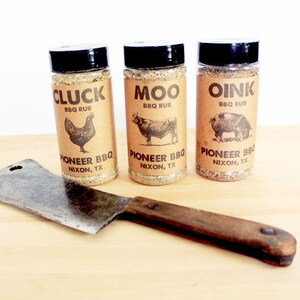 May include: Three spice rub bottles labeled "Cluck," "Moo," and "Oink" with animal illustrations, and a cleaver. The bottles are labeled "Pioneer BBQ, Nixon, TX." The cleaver has a wooden handle and a metal blade.