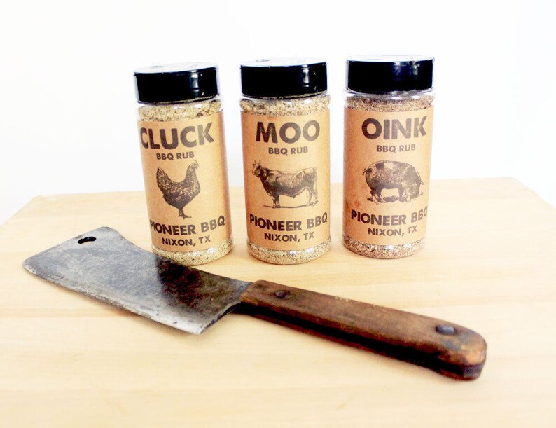 May include: Three spice rub bottles labeled "Cluck," "Moo," and "Oink" with animal illustrations, and a cleaver. The bottles are labeled "Pioneer BBQ, Nixon, TX." The cleaver has a wooden handle and a metal blade.