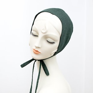 May include: A dark green bonnet with long ties, displayed on a mannequin. The bonnet is made of a soft fabric and is tied under the chin. The mannequin has closed eyes and a neutral expression.