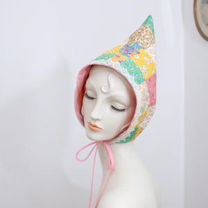 May include: A pointed bonnet featuring a patchwork design of various floral fabrics in yellow, pink, green, and white. The headwear is lined with solid pink fabric and includes thin pink ribbon ties that hang down from the chin area.