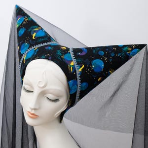 May include: A decorative hat with a celestial design, featuring a black background with blue planets and yellow accents. The hat is trimmed with a light blue and white border and is paired with a black sheer veil. The hat is displayed on a mannequin head.