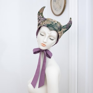 May include: A horned bonnet featuring a floral tapestry pattern in shades of green, purple, and gold. The headwear has two pointed horn-like structures at the top and a solid purple fabric ribbon tie that fastens under the chin with a bow.