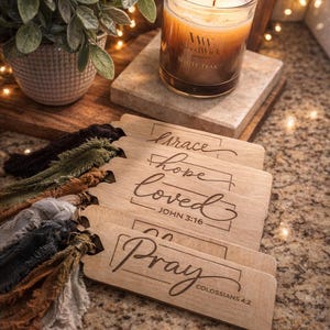 Puede incluir: Una vela encendida en un tarro de cristal con la inscripción "White Teak" descansa sobre una baldosa de mármol. Se muestran etiquetas de madera con las palabras "Grace", "Hope", "Loved" y "Pray" con borlas de colores. Una planta en maceta está en el fondo.
