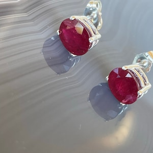 May include: A pair of silver stud earrings with oval-shaped ruby gemstones. The earrings have a simple design and are perfect for everyday wear.