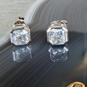 May include: A pair of silver-toned square stud earrings with a clear, faceted center stone. The earrings are set in a bezel setting, reflecting light. The earrings are displayed on a reflective surface.