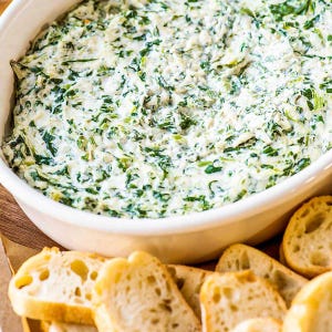Könnte beinhalten: Eine weiße ovale Schüssel gefüllt mit cremigem Spinat-Dip, einer Mischung aus grünem Spinat und einer weißen cremigen Basis. Scheiben geröstetes Brot sind vor der Schüssel angeordnet. Ein klassischer Party-Vorspeise.