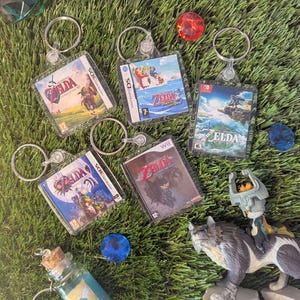 May include: Five different Zelda game cartridge keychains. Each keychain features a different Zelda game cover art, including Ocarina of Time, Twilight Princess, The Wind Waker, and Skyward Sword. The keychains are made of clear plastic and have silver key rings.