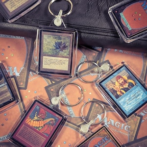 May include: A collection of clear acrylic keychains featuring miniature Magic the Gathering trading cards. Each keychain includes a metal split ring and displays a unique card illustration, including a Black Lotus, a dragon, and a wizard character.