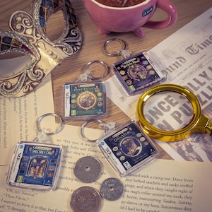 May include: A collection of four miniature Professor Layton game case keychains, a pink ceramic mug, a decorative silver and gold masquerade mask, a gold-rimmed magnifying glass, and two metal coins arranged on a printed paper background.