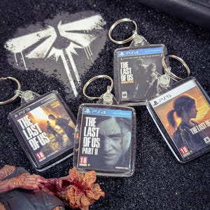 May include: Four square keychains featuring miniature game cover art for The Last of Us series on PS3, PS4, and PS5, including The Last of Us Remastered and The Last of Us Part II, alongside a small, textured, reddish-brown fungal creature figurine.