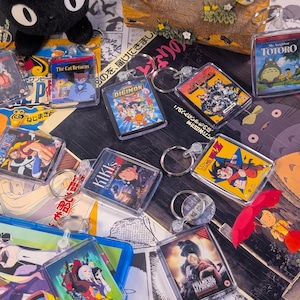 May include: A collection of square acrylic keychains featuring various anime movie and series cover art, including titles like My Neighbor Totoro, Digimon, Kiki's Delivery Service, Dragon Ball Z, Fullmetal Alchemist, and Cyberpunk Edgerunners.