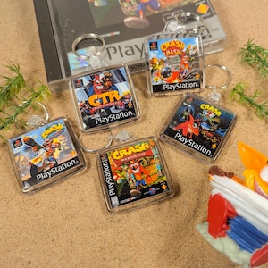 May include: Five different Crash Bandicoot video game keyrings. Each keyring is a square, clear plastic case with a printed image of a Crash Bandicoot game cover. The keyrings feature the games Crash Bandicoot, Crash Bandicoot 2: Cortex Strikes Back, Crash Bandicoot: Warped, Crash Bash, and Crash Team Racing. Each keyring has the PlayStation logo and the game title printed on it.