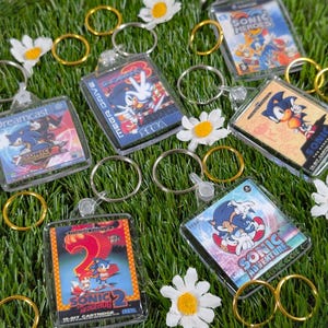 May include: A collection of square keychains featuring miniature artwork of various Sonic the Hedgehog video game covers, including Sonic Adventure, Sonic Heroes, and Sonic the Hedgehog 2, each attached to a silver metal split ring.