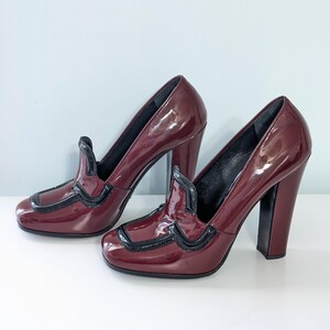 May include: A pair of burgundy patent leather high-heeled shoes with a square toe and a black trim. The heels are thick and blocky. The shoes have a classic, elegant design.