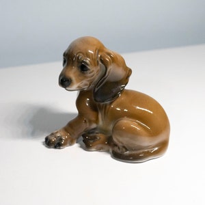 Może przedstawiać: Ceramiczna figurka  dackla w kolorze brązowym i czarnym. Figurka siedzi z głową  odwr&oacute;coną w bok i uszami  uniesionymi do g&oacute;ry.