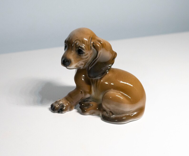 Może przedstawiać: Ceramiczna figurka  dackla w kolorze brązowym i czarnym. Figurka siedzi z głową  odwr&oacute;coną w bok i uszami  uniesionymi do g&oacute;ry.