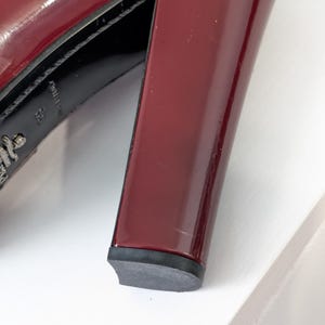 May include: Close-up of a burgundy patent leather high-heeled shoe. The shoe features a thick, block heel and a black sole. The Prada logo is visible on the sole.