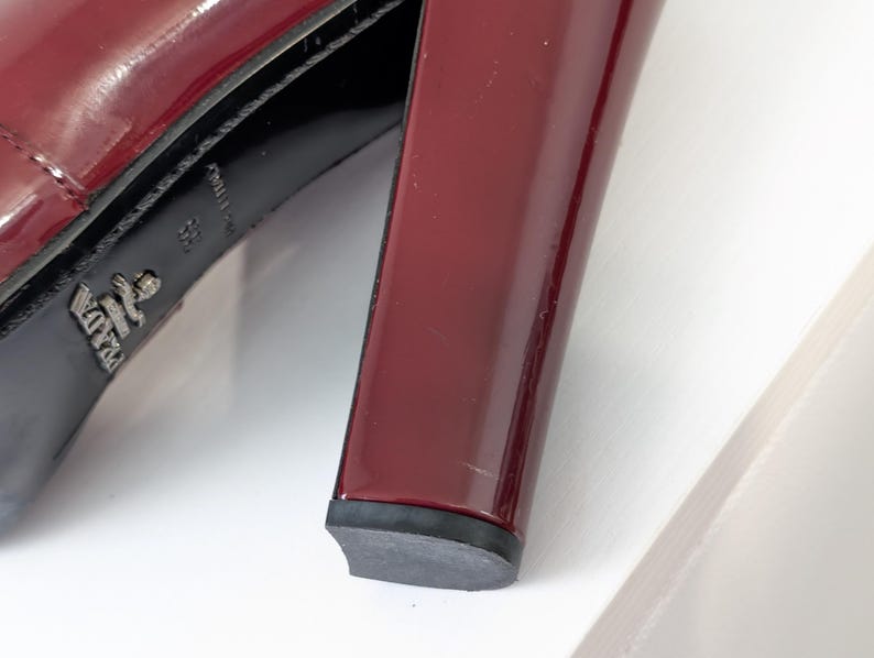 May include: Close-up of a burgundy patent leather high-heeled shoe. The shoe features a thick, block heel and a black sole. The Prada logo is visible on the sole.