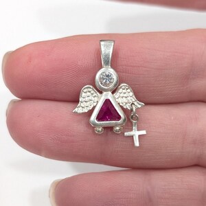 Puede incluir: Colgante de ángel de plata con una piedra triangular de color rubí y un pequeño dije de cruz. El ángel tiene alas detalladas y una piedra redonda y transparente sobre el rubí. Diseñado para ser usado como joya.