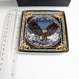 Puede incluir: Un plato decorativo cuadrado con un dise&ntilde;o detallado de &aacute;guila. El plato presenta un borde negro con detalles dorados y una imagen central de un &aacute;guila con las alas extendidas sobre un fondo monta&ntilde;oso. El plato mide aproximadamente 20 cm cuadrados.