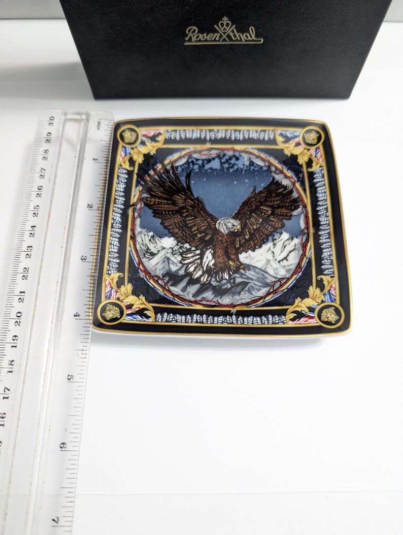 Puede incluir: Un plato decorativo cuadrado con un dise&ntilde;o detallado de &aacute;guila. El plato presenta un borde negro con detalles dorados y una imagen central de un &aacute;guila con las alas extendidas sobre un fondo monta&ntilde;oso. El plato mide aproximadamente 20 cm cuadrados.