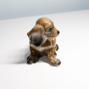 Może przedstawiać: Mała, brązowa ceramiczna figurka szczeniaka rasy dachshund siedzącego z głową odchyloną na bok. Figurka ma błyszczące wykończenie i jest szczeg&oacute;łowo wykonana z czarnymi oczami i nosem.