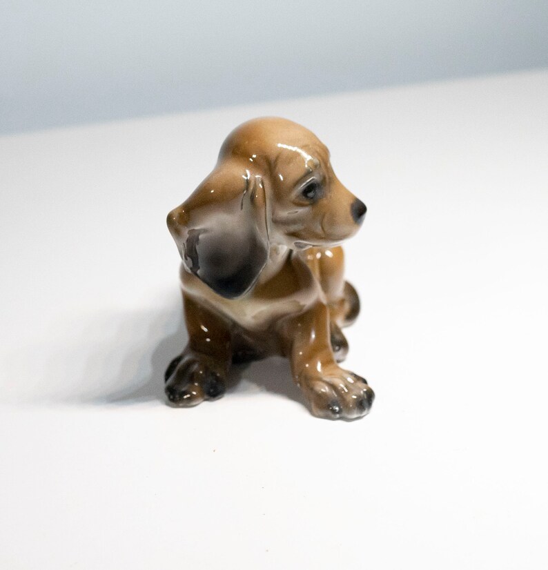 Może przedstawiać: Mała, brązowa ceramiczna figurka szczeniaka rasy dachshund siedzącego z głową odchyloną na bok. Figurka ma błyszczące wykończenie i jest szczeg&oacute;łowo wykonana z czarnymi oczami i nosem.