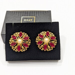 May include: A pair of gold tone earrings with a red flower design and a pearl center. The earrings are in a black velvet box with the word "AVON" printed on the box.